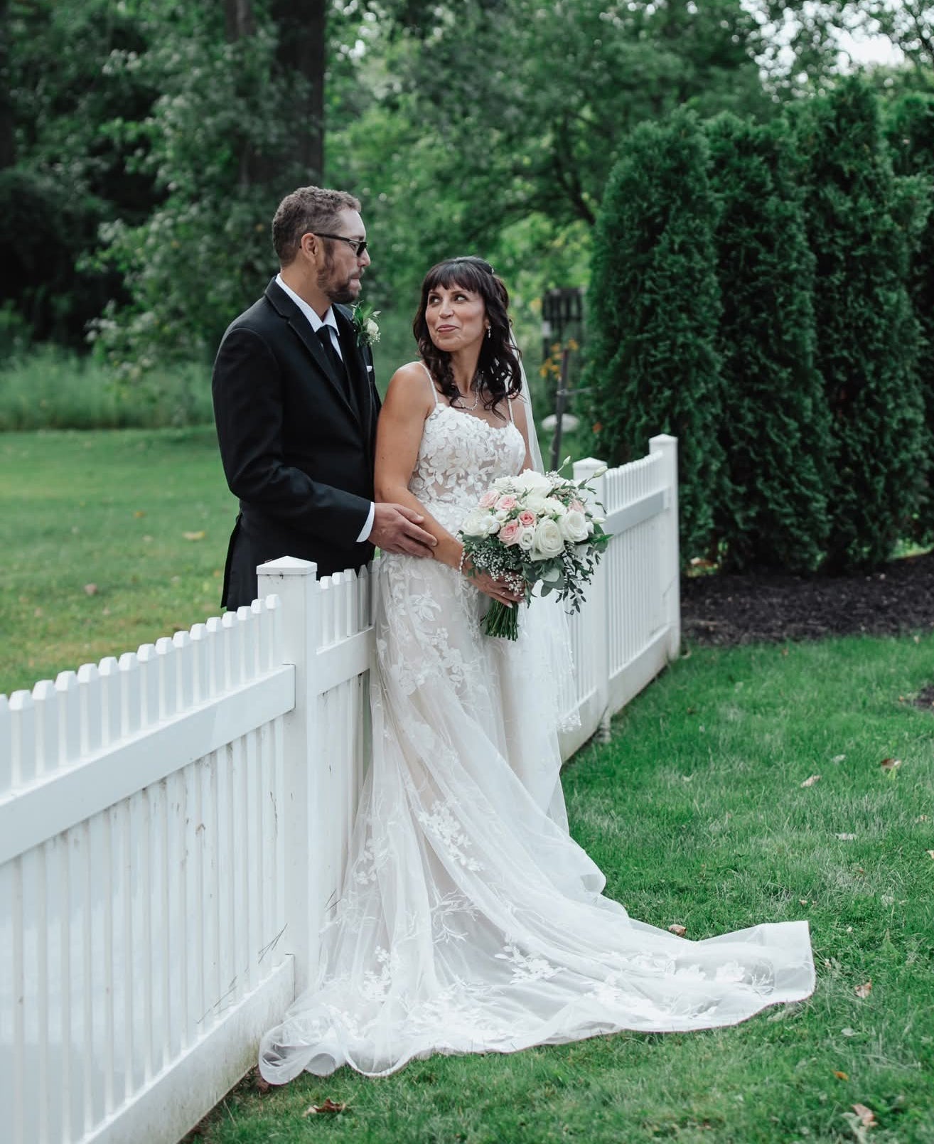 Wedding couple standing by a white picket fence with greenery in the background